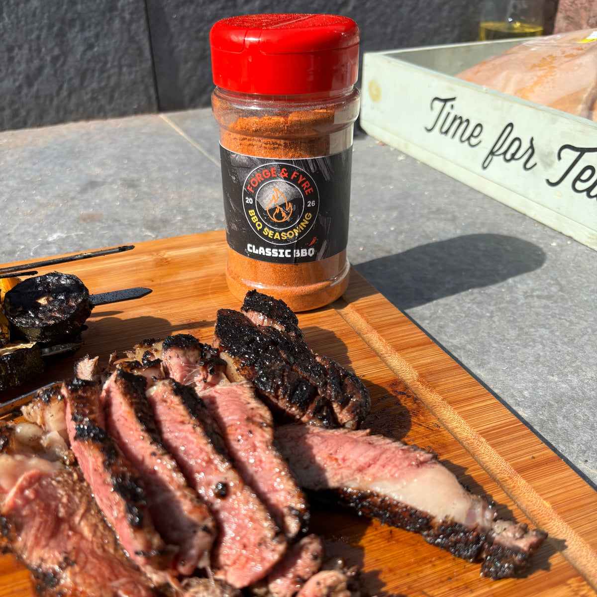 Thick slices of medium-rare steak with a dark peppery crust on a wooden board, a jar of Forge & Fyre Classic BBQ rub behind