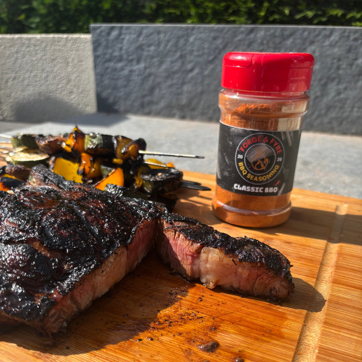A jar of Forge & Fyre Classic BBQ rub on a wooden board beside a sliced char-crusted steak and grilled vegetable skewers
