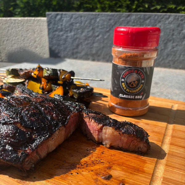 A jar of Forge & Fyre Classic BBQ rub on a wooden board beside a sliced char-crusted steak and grilled vegetable skewers