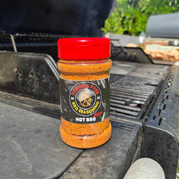 A bottle of Hot BBQ spice rub by Forge & Fyre BBQ Seasoning sits on the edge of a black grill outdoors