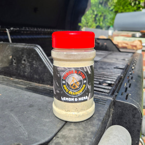 A bottle of Lemon & Herb BBQ spice rub by Forge & Fyre BBQ Seasoning sits on the edge of a black grill outdoors