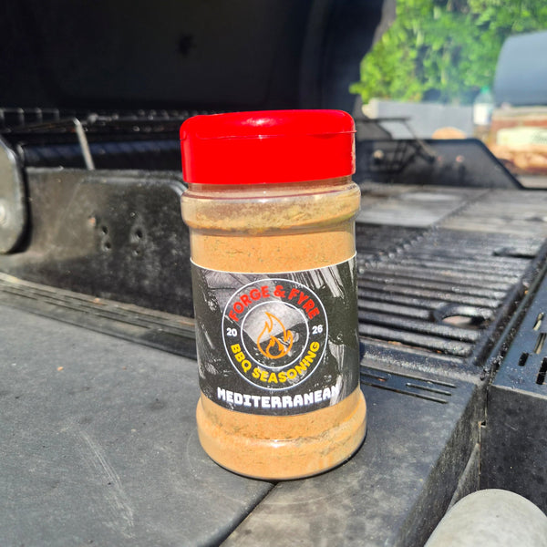 A bottle of Mediterranean Style BBQ spice rub by Forge & Fyre BBQ Seasoning sits on the edge of a black grill outdoors