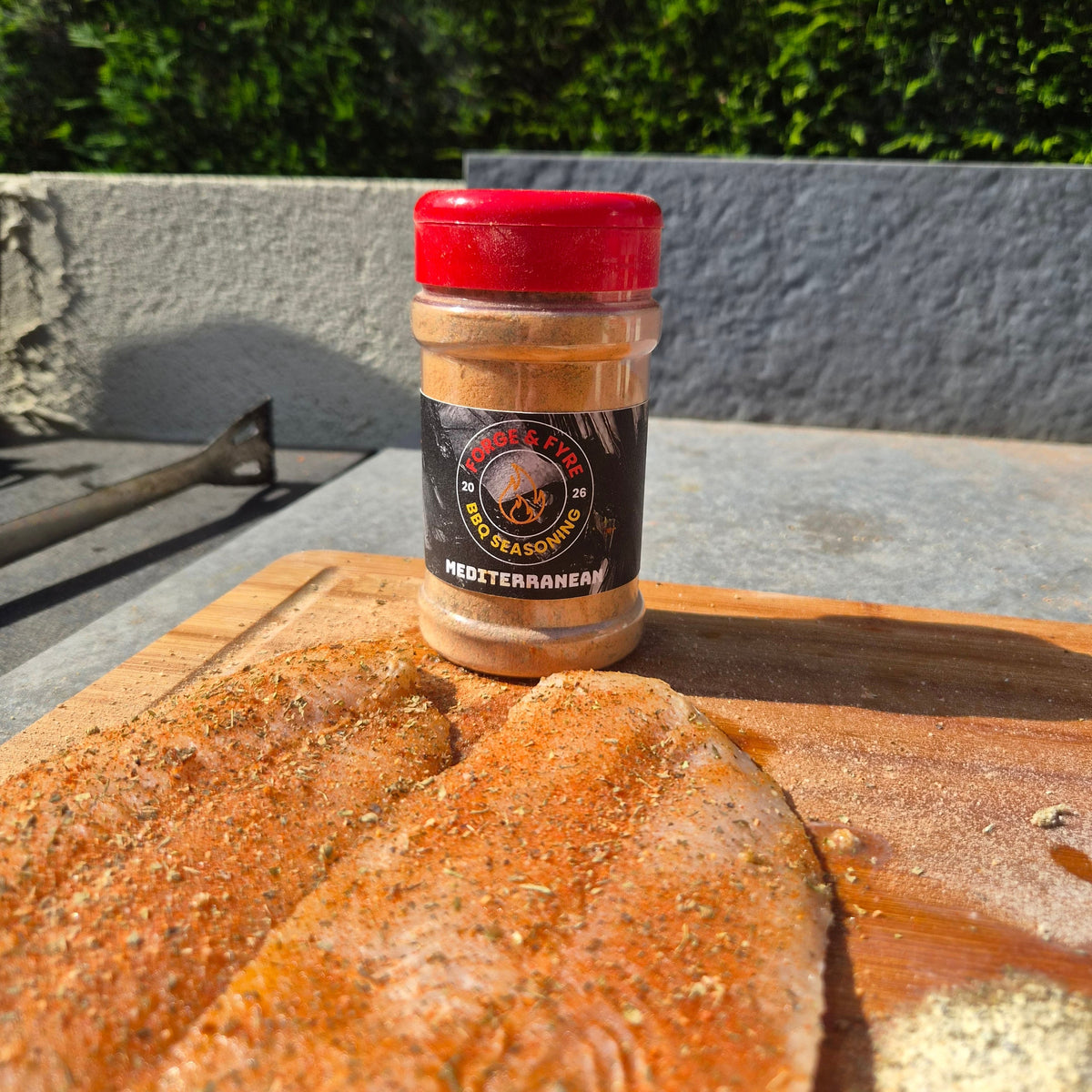 Two fish fillets coated in Forge & Fyre Mediterranean rub on a wooden board, with the jar of seasoning alongside