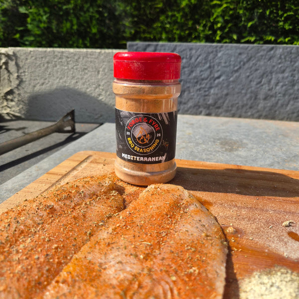 Two fish fillets coated in Forge & Fyre Mediterranean rub on a wooden board, with the jar of seasoning alongside