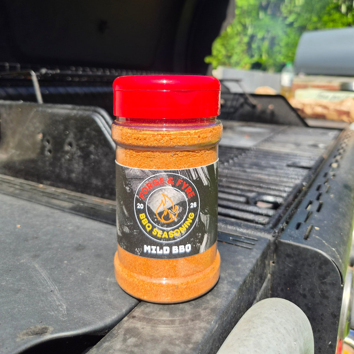 A bottle of Mild BBQ spice rub by Forge & Fyre BBQ Seasoning sits on the edge of a black grill outdoors