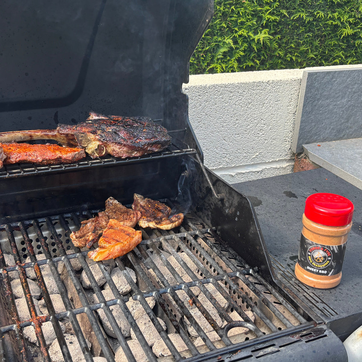 Ribs and steak searing on a black barbecue grill with a jar of Forge & Fyre Sweet BBQ rub in the foreground