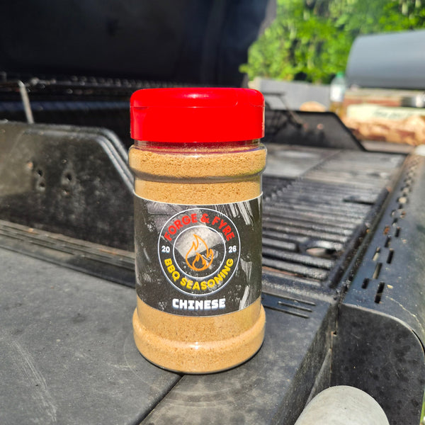 A bottle of Chinese Style BBQ spice rub by Forge & Fyre BBQ Seasoning sits on the edge of a black grill outdoors