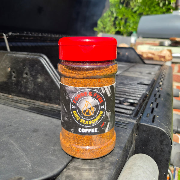A bottle of Coffee BBQ spice rub by Forge & Fyre BBQ Seasoning sits on the edge of a black grill outdoors