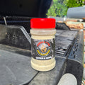 A bottle of Lemon & Herb BBQ spice rub by Forge & Fyre BBQ Seasoning sits on the edge of a black grill outdoors