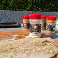 Jar of Lemon & Herb seasoning by Forge & Fyre BBQ Seasonings is arranged on a wooden cutting board outdoors, with a seasoned fish fillet in the foreground
