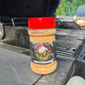 A bottle of Mediterranean Style BBQ spice rub by Forge & Fyre BBQ Seasoning sits on the edge of a black grill outdoors