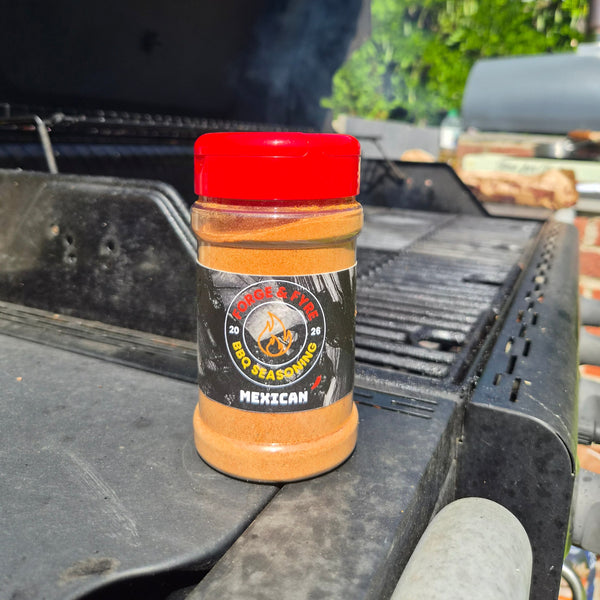 A bottle of Mexican Style BBQ spice rub by Forge & Fyre BBQ Seasoning sits on the edge of a black grill outdoors