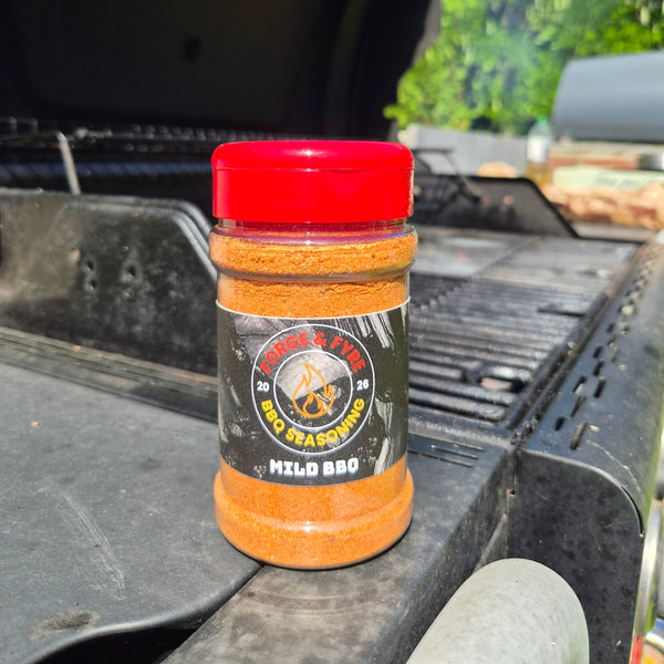A bottle of Mild BBQ spice rub by Forge & Fyre BBQ Seasoning sits on the edge of a black grill outdoors