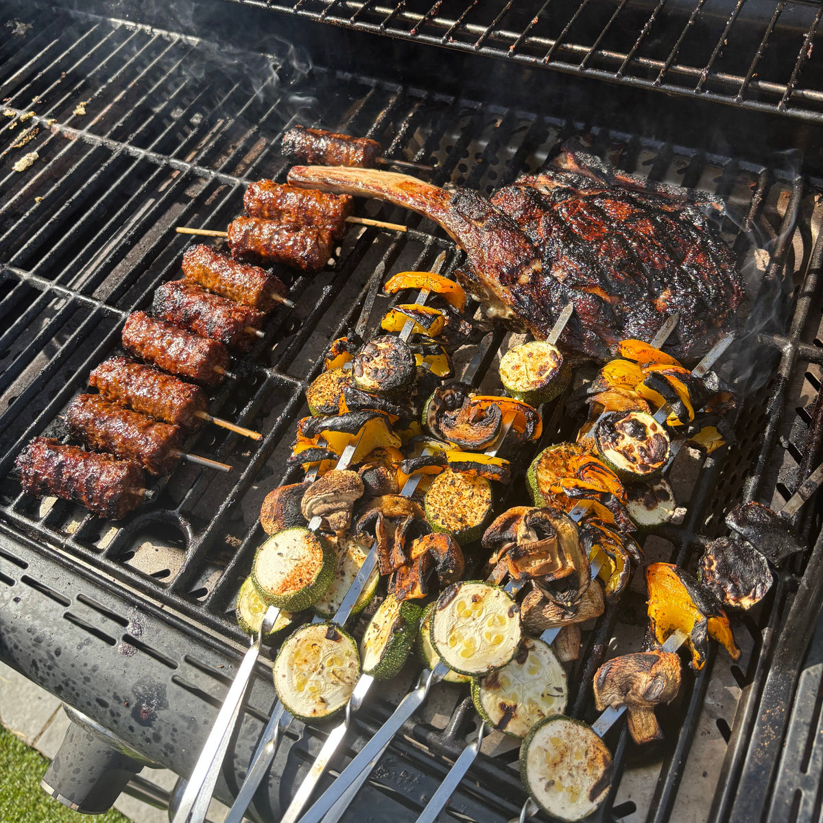 Vegetable skewers and a tomahawk steak cooking over the bars of a black barbecue grill