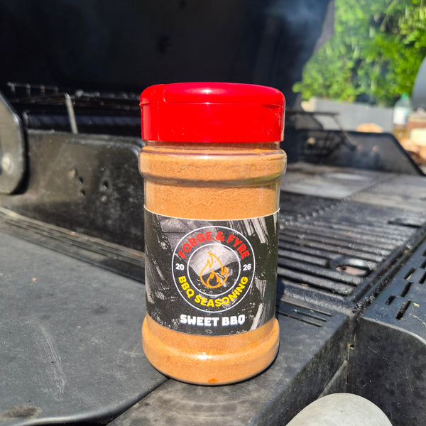 A bottle of Sweet BBQ spice rub by Forge & Fyre BBQ Seasoning sits on the edge of a black grill outdoors