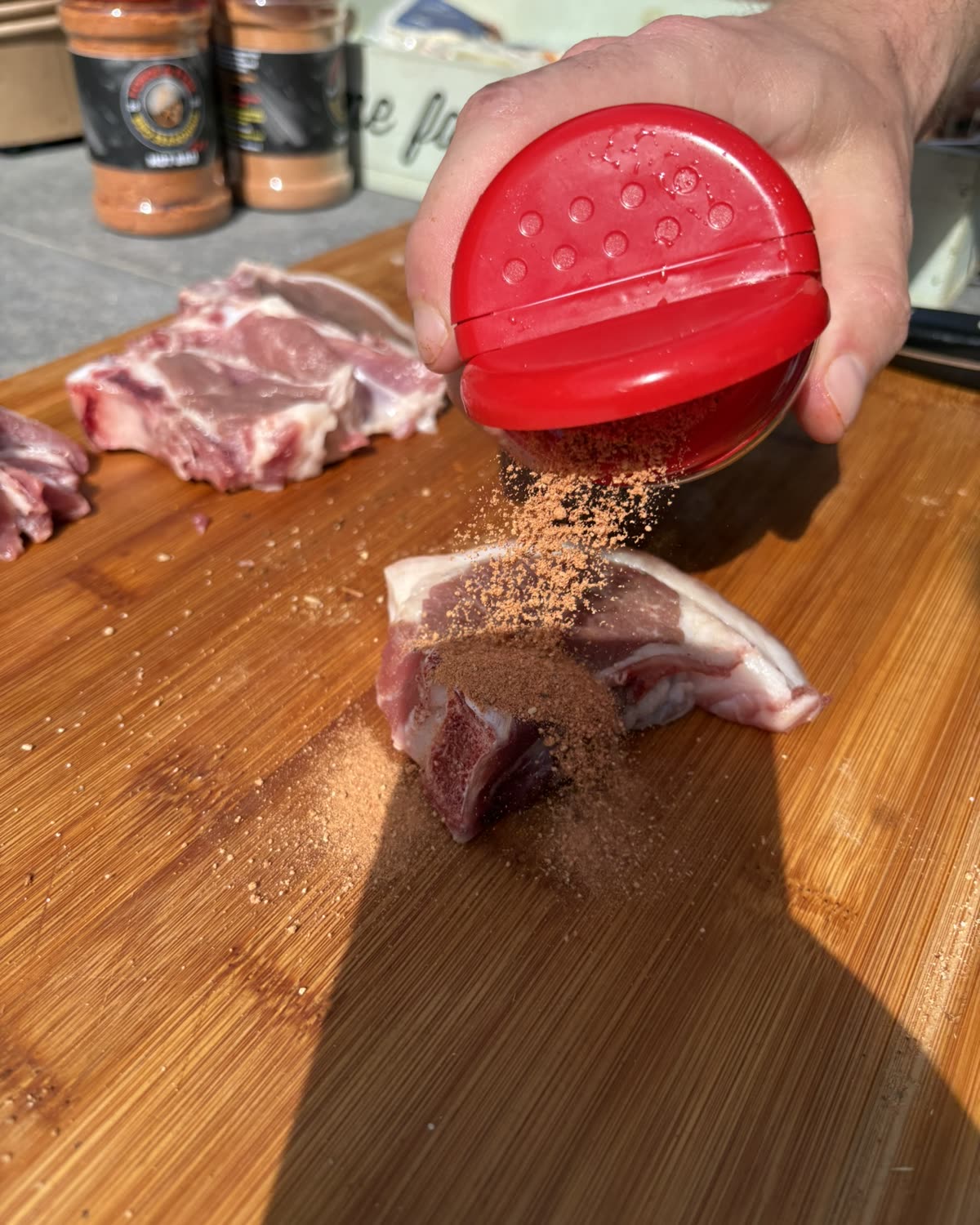 A hand shaking Forge & Fyre seasoning over lamb chops on a wooden board
