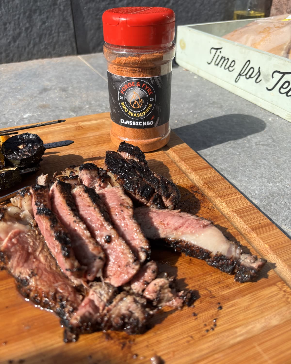 Sliced tomahawk steak on a board next to a jar of Forge & Fyre Classic BBQ seasoning